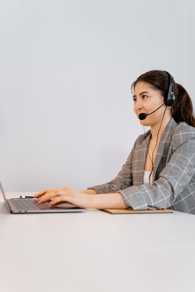 Professional woman in plaid blazer using laptop with headset in an office setting.